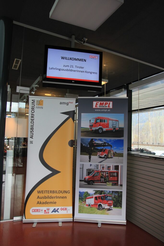 Bild von 2 Roll-Ups und einem Bildschirm mit der Anzeige "Willkommen zum 21. Tiroler Lehrlingsausbilder*innen Kongress"
