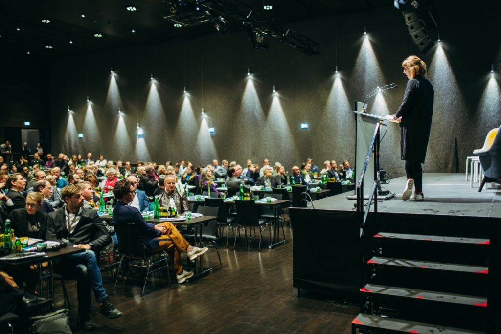 Bild von der Bühne und vom Publikum beim Kongress. Auf der Bühne hält eine Frau eine Präsentation.