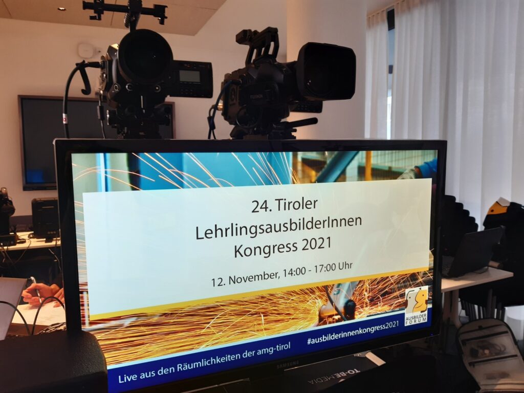 Bild von einem großen Bildschirm mit der Anzeige "24. Tiroler Lehrlingsausbilder*innen Kongress 2021", im Hintergrund zwei Kameras auf Stativen
